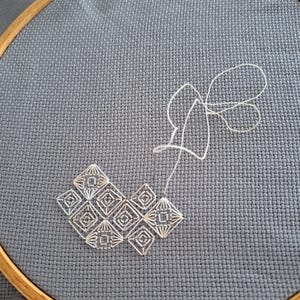 May include: A close-up of a cross-stitch embroidery project in progress. The design features a geometric pattern in white thread on a grey fabric stretched within a wooden hoop. The needle and thread are visible, showing the stitching process.