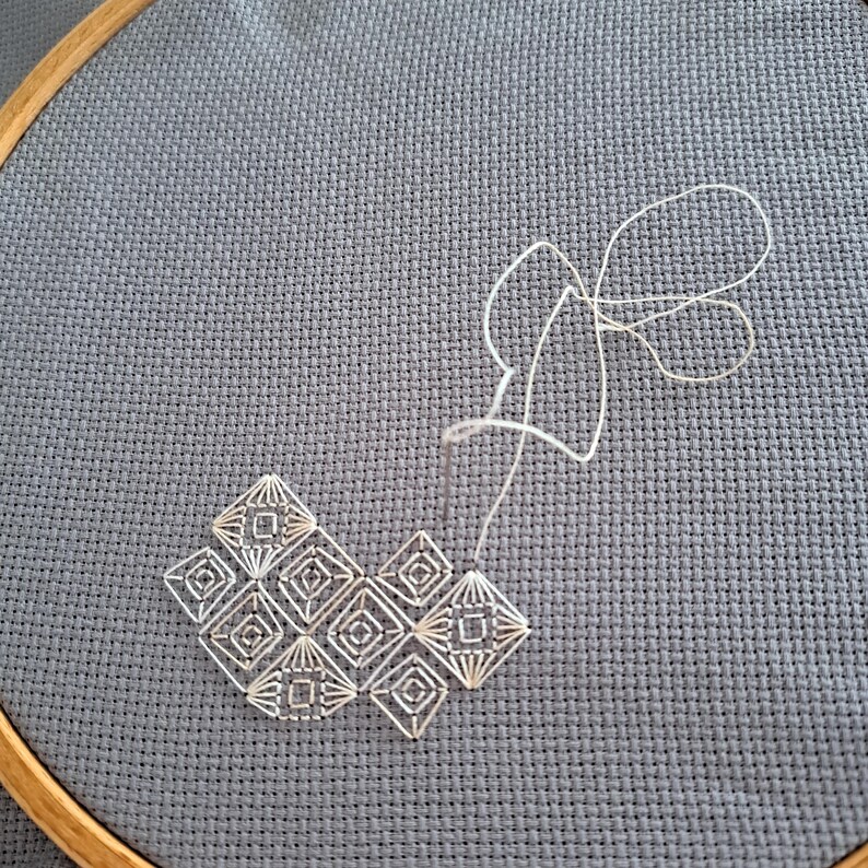 May include: A close-up of a cross-stitch embroidery project in progress. The design features a geometric pattern in white thread on a grey fabric stretched within a wooden hoop. The needle and thread are visible, showing the stitching process.