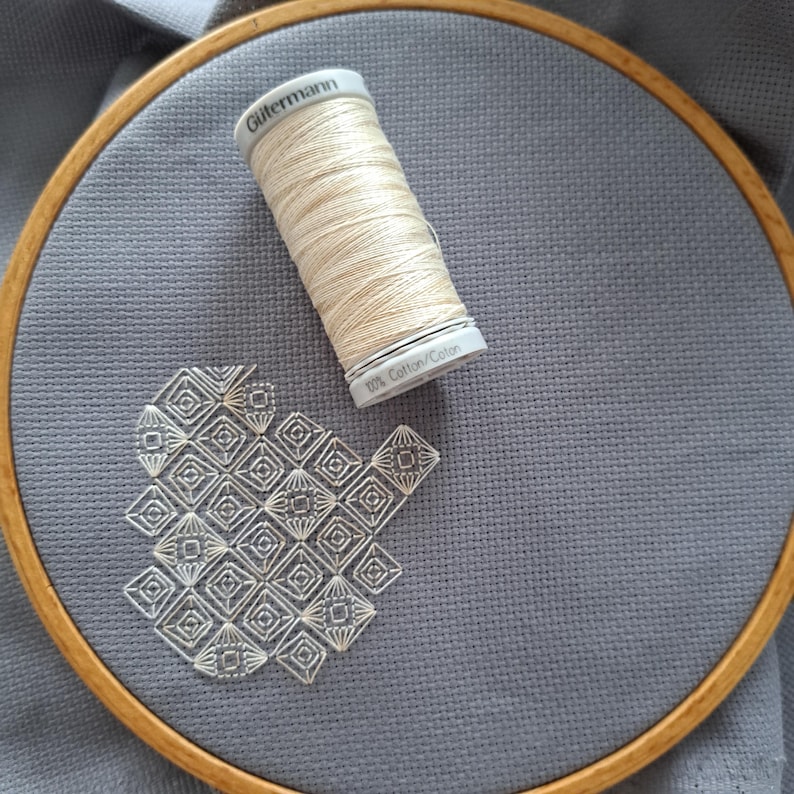 May include: A grey fabric stretched within a wooden embroidery hoop, showcasing a geometric design stitched with cream-coloured thread. A spool of G&uuml;termann cream-coloured thread sits on the fabric. The thread spool is labelled "100% Cotton/Coton".