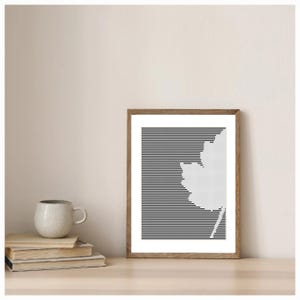 Könnte beinhalten: Ein gerahmter Kunstdruck mit einem weißen Blattdesign vor einem Hintergrund aus schwarzen horizontalen Linien. Das Kunstwerk befindet sich in einem Holzrahmen und wird auf einer hellen Oberfläche präsentiert. Ein Stapel Bücher und eine Keramiktasse befinden sich ebenfalls auf der Oberfläche.