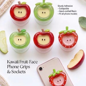 May include: A collection of six resin phone grips shaped like apples and strawberries with faces. The grips feature red and green colors, stems, and leaves. One strawberry grip is attached to a smartphone. Text reads: Kawaii Fruit-Face Phone Grips & Sockets, Sturdy Adhesive, Collapsible, Hand-crafted Resin, Fit all phone models.
