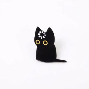 May include: A black enamel pin shaped like a sitting cat with large yellow eyes and a white loading icon symbol centered on its forehead. The cat has pointed ears and a long tail extending to the right, rendered in a smooth, solid black finish.