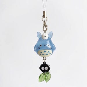 May include: A blue and white creature charm with a small white figure on top, suspended from a silver-toned metal lobster clasp and white cord. Below the main figure hangs a black spiky soot sprite charm with two translucent green leaves.
