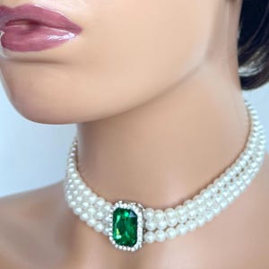 Puede incluir: Un collar gargantilla de perlas blancas con tres filas de perlas y una gran piedra preciosa verde esmeralda en el centro.