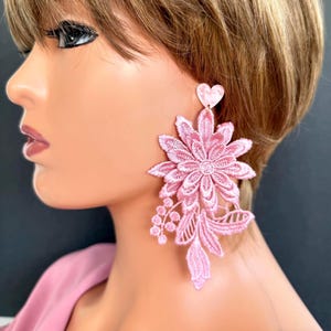 May include: Pink floral lace earrings with a heart-shaped stud. The earrings feature a large, detailed flower design with delicate leaves and berry-like accents. The earrings are a soft, pastel pink color.