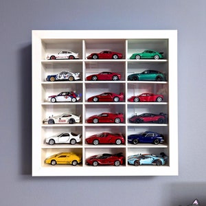 May include: A white display case filled with miniature toy cars in various colours, including red, yellow, white, and blue. The case is mounted on a grey wall. Text at the bottom reads "*IKEA Sannähed frame not included."