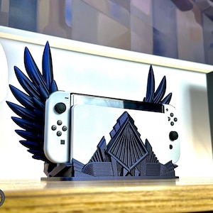 Può includere: Una console Nintendo Switch bianca e nera è esposta in un supporto a forma di ala blu scuro. Il supporto ha un design geometrico alla base. L'immagine mostra anche una statuina di gatto nero e un gatto dei cartoni animati che tiene un cartello.