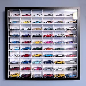 Può includere: Una vetrina nera piena di piccole macchinine giocattolo di vari colori. Le auto sono disposte su ripiani bianchi. La vetrina è montata su una parete azzurra. Il testo "*IKEA Sannahed frame not included" è in basso.