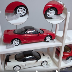 Pu&ograve; includere: Uno scaffale espositivo in plastica bianca per modellini di auto con bloccaruote semitrasparenti. Il testo riporta: Secure Wheel-Locks, finished in discreet semi-clear, use them flat or angled for extra hold, Easy snap-together assembly, Integrated wheel-locking design, e Verified fit for: Mini GT, Tarmac Works, Matchbox, Tomica & More!