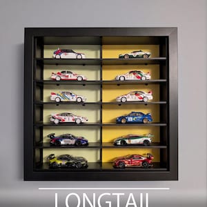 Pu&ograve; includere: Una vetrinetta da parete nera con 12 scomparti per modellini di auto. L'interno presenta pannelli posteriori che si alternano tra verde chiaro e giallo. Il testo in basso recita: LONGTAIL BLACK 12 SLOTS | LONG CARS | UP TO 90mm.