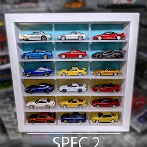 Pu&ograve; includere: Una vetrinetta bianca con 18 scomparti contenente varie macchinine giocattolo in miniatura. L'unit&agrave; presenta un design aperto e un pannello posteriore azzurro e giallo. Il testo in basso recita: SPEC.2, SEMI-CLEAR, 18 SLOTS | OPEN DESIGN | UP TO 70mm.