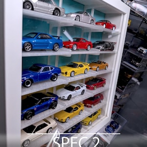 Pu&ograve; includere: Una vetrinetta da parete bianca con pannello posteriore azzurro, dotata di 18 scomparti per modellini di auto. La vetrina ha un design aperto e pu&ograve; ospitare modelli fino a 70mm. Il testo riporta: SPEC.2, SEMI-CLEAR, 18 SLOTS, OPEN DESIGN, UP TO 70mm.