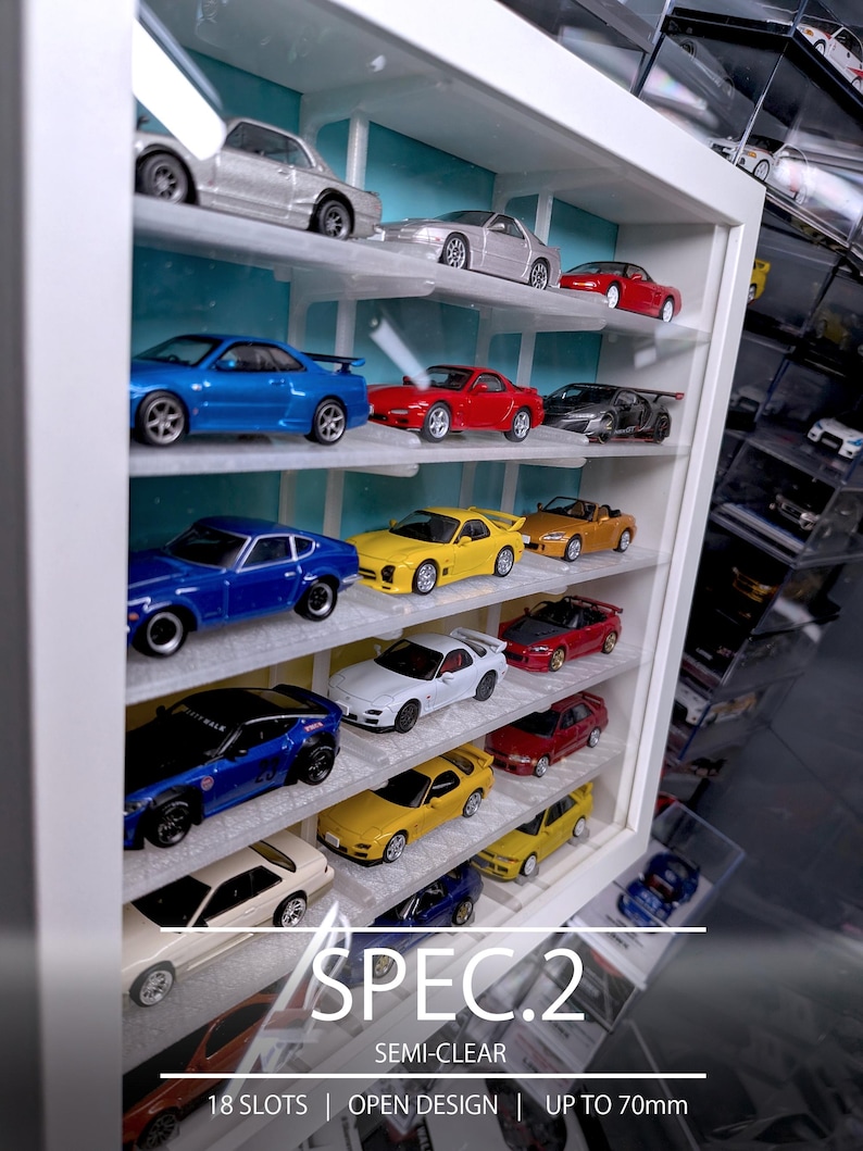Pu&ograve; includere: Una vetrinetta da parete bianca con pannello posteriore azzurro, dotata di 18 scomparti per modellini di auto. La vetrina ha un design aperto e pu&ograve; ospitare modelli fino a 70mm. Il testo riporta: SPEC.2, SEMI-CLEAR, 18 SLOTS, OPEN DESIGN, UP TO 70mm.