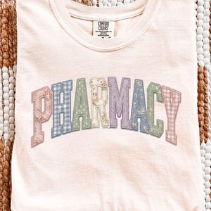May include: An ivory short-sleeve t-shirt with a crew neckline and the word PHARMACY printed in an arched, collegiate-style font. Each letter features a unique pastel-colored pattern, including floral, gingham, and polka dot designs.