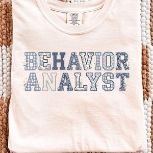May include: An ivory short-sleeve t-shirt with a crew neckline. The front features the text BEHAVIOR ANALYST in block letters. Each letter has a unique blue and white pattern, including gingham, floral, and solid designs. A Comfort Colors label is visible.
