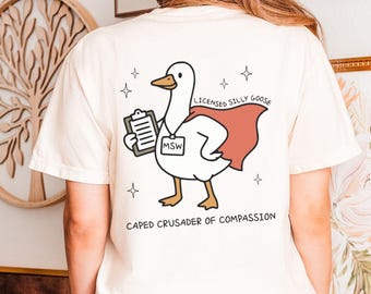 May include: Ivory-colored t-shirt featuring a cartoon goose wearing a red cape and holding a clipboard. The text on the shirt reads "LICENSED SILLY GOOSE" and "CAPED CRUSADER OF COMPASSION". The design includes stars.