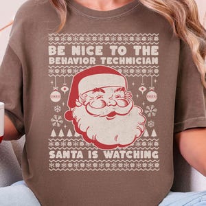 May include: A brown t-shirt with the text "BE NICE TO THE BEHAVIOR TECHNICIAN SANTA IS WATCHING" and a Santa Claus graphic. The person is holding a white and red mug with a gingerbread man design.