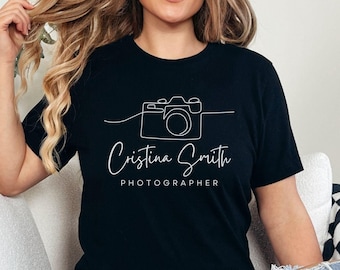 May include: Black t-shirt with a white line drawing of a camera and the text "Cristina Smith PHOTOGRAPHER". The shirt is worn by a person sitting on a white couch. The person is wearing blue jeans.