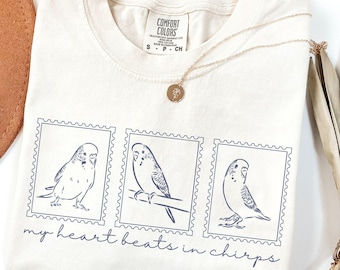 以下が含まれることがあります： 丸首のアイボリー色のTシャツ。青色のインクで描かれた、切手フレームの中のセキセイインコのイラストが3つあります。「my heart beats in chirps」という文字が下に印刷されています。金のネックレスとペンダントがシャツの上にあります。