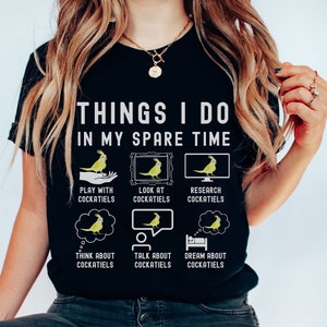 May include: A black t-shirt with the text "Things I do in my spare time" and illustrations of cockatiels. The illustrations show a cockatiel playing, looking at a picture, researching on a computer, thinking, talking, and sleeping.