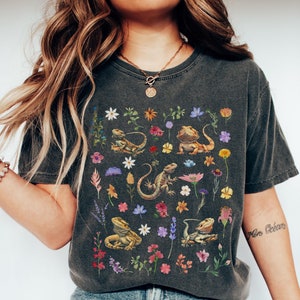 Peut inclure: Un t-shirt gris foncé avec un imprimé floral représentant plusieurs dragons barbus et des fleurs. Le t-shirt a un col rond et des manches courtes.
