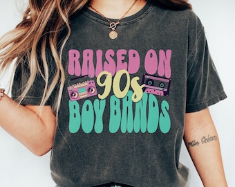 May include: A woman wearing a charcoal grey t-shirt with the text "RAISED ON 90s BOY BANDS" in pink, yellow, and teal. The shirt features a retro boombox and cassette tape graphic.