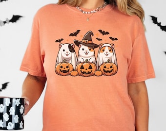May include: A peach-coloured t-shirt featuring a graphic of three guinea pigs dressed as ghosts for Halloween. The guinea pigs are wearing witch hats and holding pumpkins. The shirt has a crew neck and short sleeves.