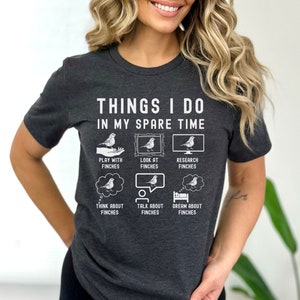 May include: A dark grey t-shirt with a white graphic that says "Things I do in my spare time" and shows a series of illustrations of finches. The illustrations show a person playing with a finch, looking at a finch in a picture frame, researching finches on a computer, thinking about finches, talking about finches, and dreaming about finches.