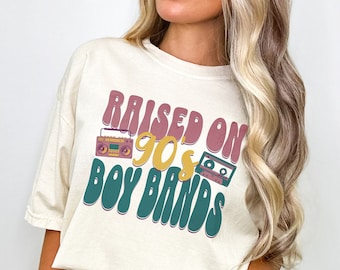 May include: A woman wearing a cream colored t-shirt with the text "RAISED ON 90's BOY BANDS" in a retro font. The shirt features a vintage cassette tape and boombox graphic.
