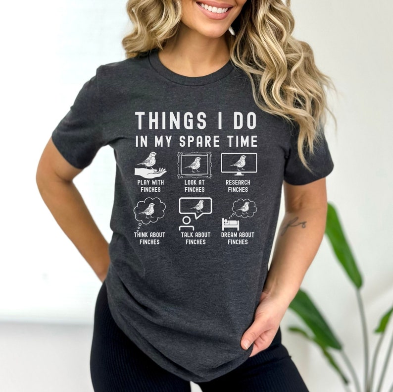 May include: A dark grey t-shirt with a white graphic that says "Things I do in my spare time" and shows a series of illustrations of finches. The illustrations show a person playing with a finch, looking at a finch in a picture frame, researching finches on a computer, thinking about finches, talking about finches, and dreaming about finches.