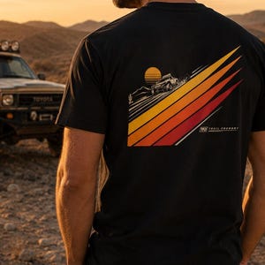 May include: Black t-shirt featuring a vehicle graphic driving up a diagonal striped design in orange, yellow, and red, with a setting sun. The shirt is worn by a person wearing a black cap and sunglasses, standing in a desert landscape.