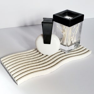 May include: A white ceramic soap dish with a wavy design and black accents. A white round soap and a clear glass jar with a black lid filled with white cotton swabs are on the dish.