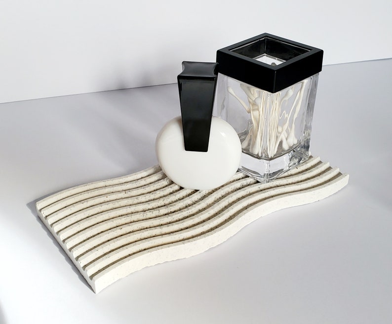 May include: A white ceramic soap dish with a wavy design and black accents. A white round soap and a clear glass jar with a black lid filled with white cotton swabs are on the dish.