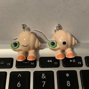 May include: A pair of dangle earrings featuring a small, light pink, 3D creature with green eyes and orange feet. The creature has a small mouth and a slightly surprised expression.