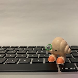May include: A small, beige, cartoon-like figure with a green eye and pink feet stands on a black keyboard. The figure is holding a small, green object in its hand.