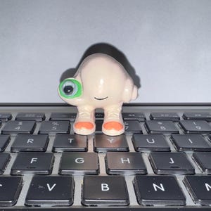 May include: A small, pale, rounded figurine with a single large green-rimmed eye, a simple black line for a mouth, and two legs wearing orange-toed shoes. The character has a smooth, light-colored surface and stands upright on a black keyboard.