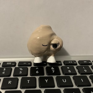 May include: A small, beige, 3D printed figurine with a single eye and a closed-mouth smile. The figurine is standing on a keyboard.