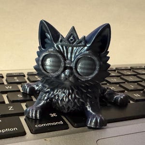May include: A dark blue cat figurine with large, round goggles and a crown, resting on a laptop keyboard. The cat has a textured surface and is posed with its paws extended. The keyboard keys are black.