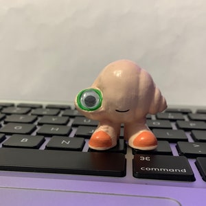 May include: A small, pink, hand-painted figurine with a green eye and orange feet. The figurine is standing on a black keyboard.