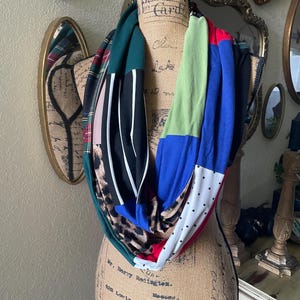 Puede incluir: Bufanda infinita de patchwork con una variedad de patrones y colores, incluyendo estampado de leopardo, rayas, lunares y bloques sólidos de verde, azul y rojo. La bufanda se exhibe en un maniquí.