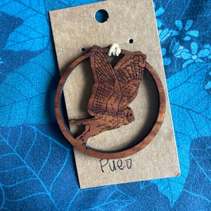 May include: A wooden owl pendant with a circular frame, laser-cut with intricate details. The owl is perched on a branch with its wings spread. The word "Pueo" is written in cursive on the attached tag. The pendant is attached to a brown tag with a hole.