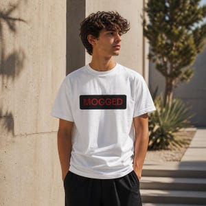 Könnte beinhalten: Weißes T-Shirt mit kurzen Ärmeln und dem Wort "MOGGED" in Rot in einem schwarzen Rechteck. Das T-Shirt ist aus weichem Material. Die Person trägt schwarze Shorts.