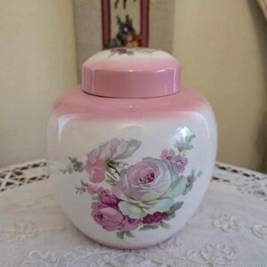 May include: A white and pink ceramic jar with a lid, featuring a floral rose design. The jar has a rounded shape and is decorated with pink roses and green leaves. The lid is also pink.