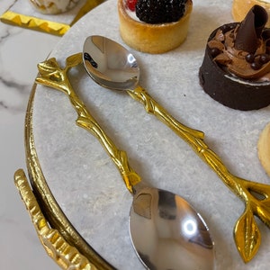 Puede incluir: Dos cucharas doradas con mangos en forma de hoja y cuencos plateados. Las cucharas están sobre una superficie de mármol blanco con un borde dorado.