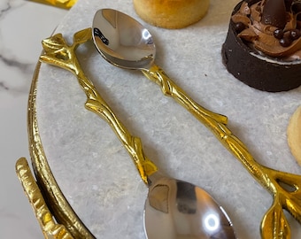 May include: Two gold-tone spoons with leaf-shaped handles and silver-tone bowls. The spoons are on a white marble surface with a gold rim.
