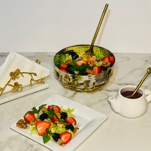 May include: A white ceramic teapot with a gold-colored twig-shaped spoon inside, filled with a dark red liquid. A white marble napkin holder with a gold-colored twig design holds a white napkin. A white square plate with a salad of strawberries, blackberries, walnuts, and lettuce. A gold-colored bowl with a hammered finish holds a salad of strawberries, blackberries, walnuts, and lettuce. A gold-colored twig-shaped spoon is in the bowl.