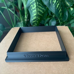 May include: A black plastic square frame with the text "12cm x 12cm" printed on the bottom edge. The frame is sitting on a brown surface.