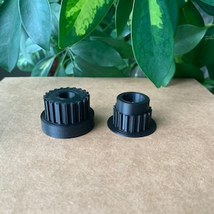 May include: Two black plastic gears with teeth, one larger than the other, on a brown surface.