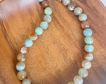 Puede incluir: Un collar de cuentas redondas y facetadas en tonos azules, marrones y crema. El collar tiene un cierre de color cobre y se exhibe en una bandeja de madera. Las cuentas tienen aproximadamente 1,3 cm de diámetro.