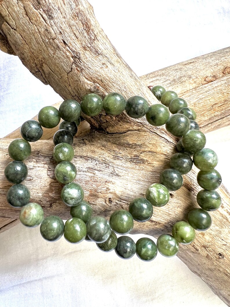 Puede incluir: Dos pulseras de cuentas de jade verde con un patr&oacute;n moteado verde y blanco. Las pulseras est&aacute;n apiladas una encima de la otra y descansan sobre un trozo de madera flotante.